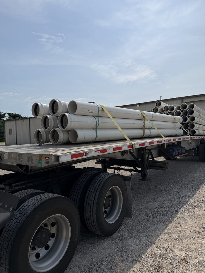 Najax Trucking flatbed trailer loaded with white PVC pipe, secured with yellow straps under a blue sky